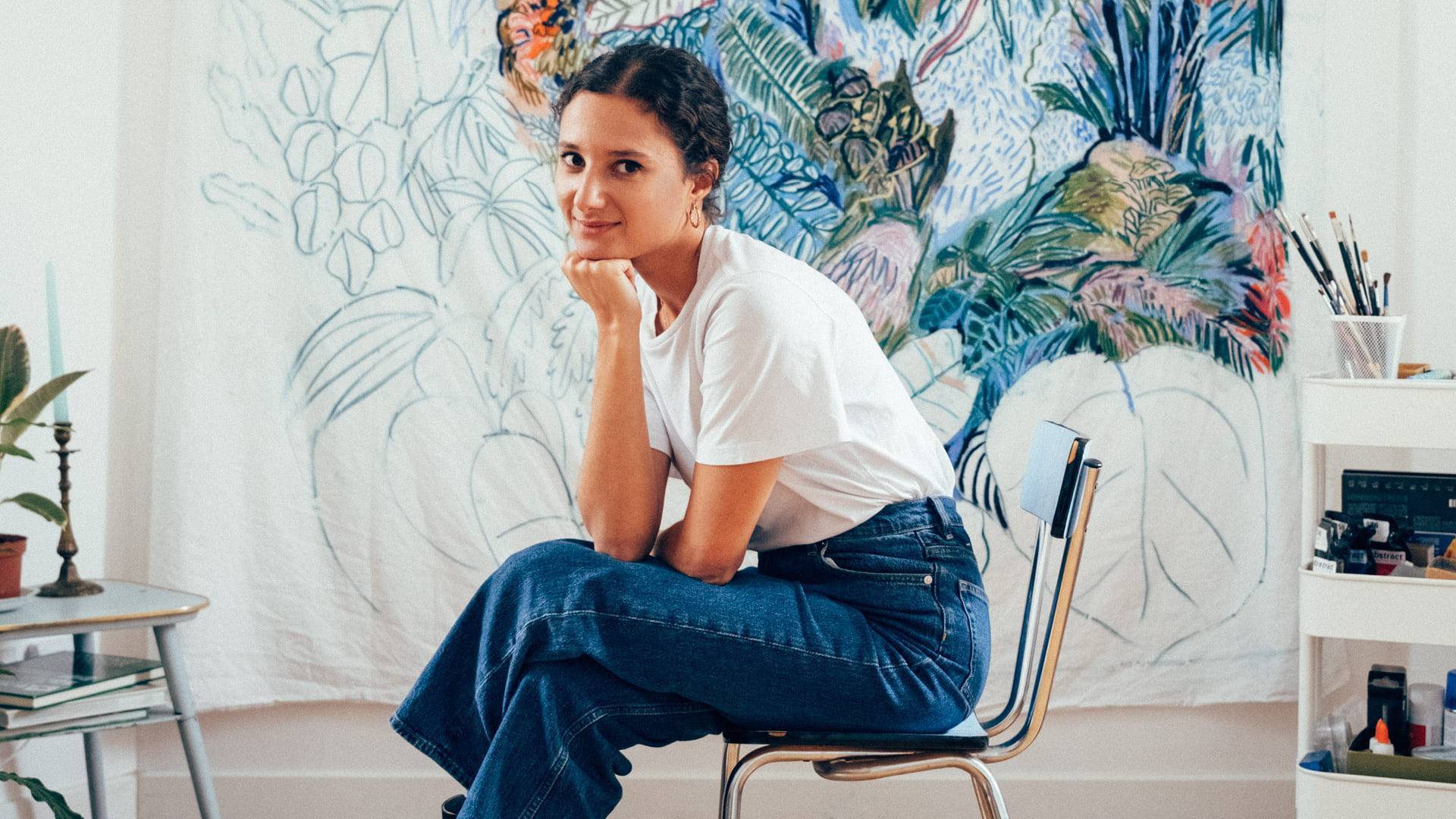 photo de Beya Rebai assise sur une chaise dans un atelier avec un dessin en arrière plan